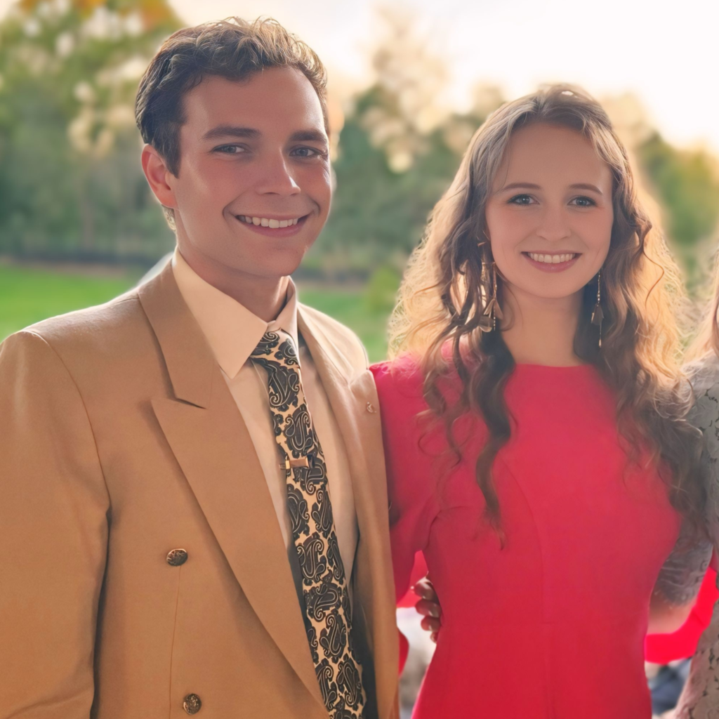 A man with curly light brown hair in a tan suit jacket standing next to a woman with long curly brown hair wearing a bright pink dress